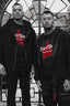 Two men wearing black Dirtbag Mega Zip Hoodies layered over Dirtbag Attitude T Shirts with bold red DBA star logos, standing in front of an iron gate in a gritty urban setting. The image highlights the oversized zipper detail on the Mega Zip Hoodies, alternative streetwear style, attitude driven apparel and the bold Dirtbag lifestyle aesthetic.