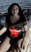 Woman sitting on a boat at sea wearing a black Dirtbag Attitude T Shirt with a bold red DBA star logo, enjoying an ocean backdrop in bright sunlight. The image highlights alternative streetwear style, coastal adventure lifestyle clothing, graphic tees for women, and the bold attitude driven Dirtbag apparel aesthetic.